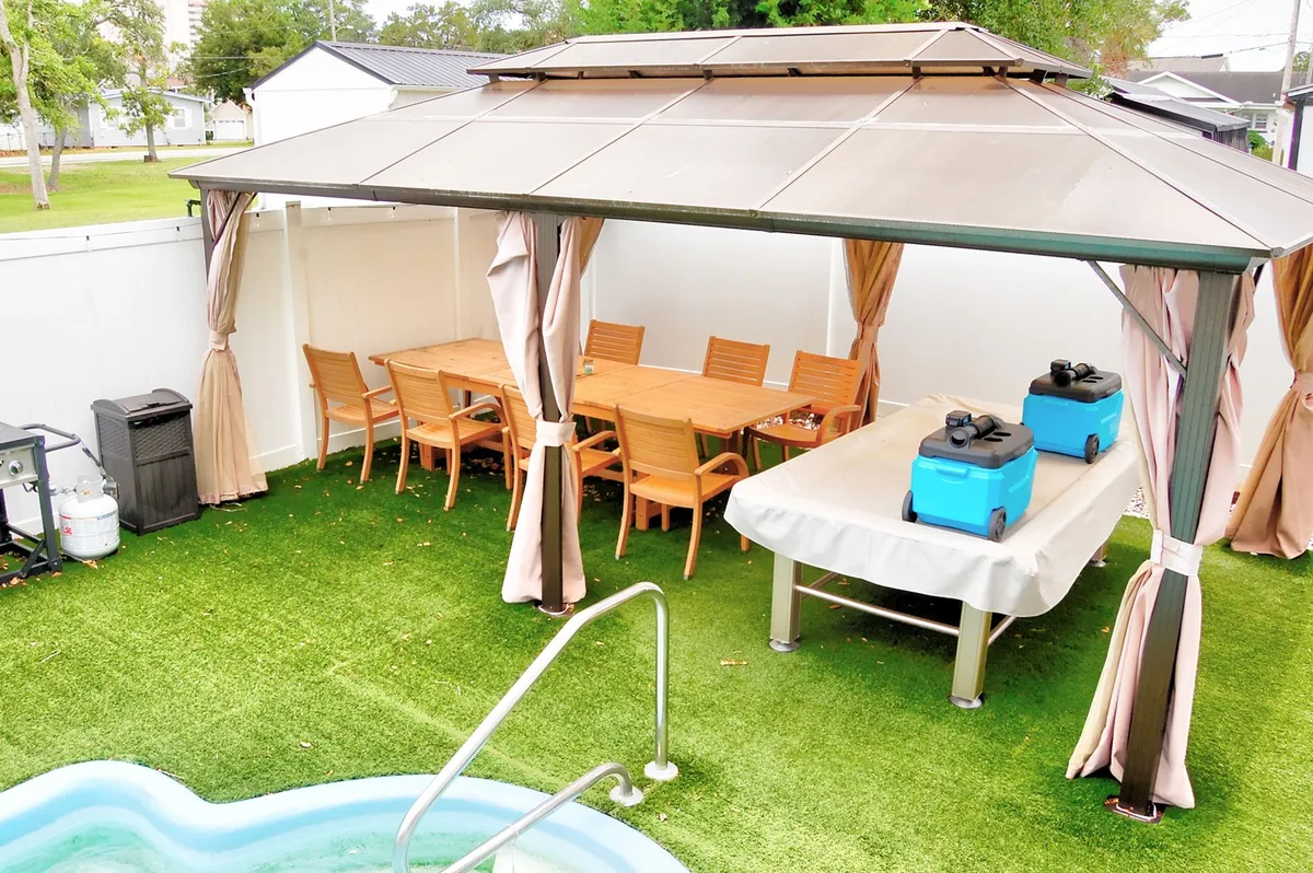 An inviting outdoor dining area set under a gazebo, with a large table for eight, a barbecue grill, and a view of the adjacent swimming pool. The setup is ready for lively outdoor gatherings, summer barbecues, or starlit feasts, complete with convenient access to grilling and chilled beverages.