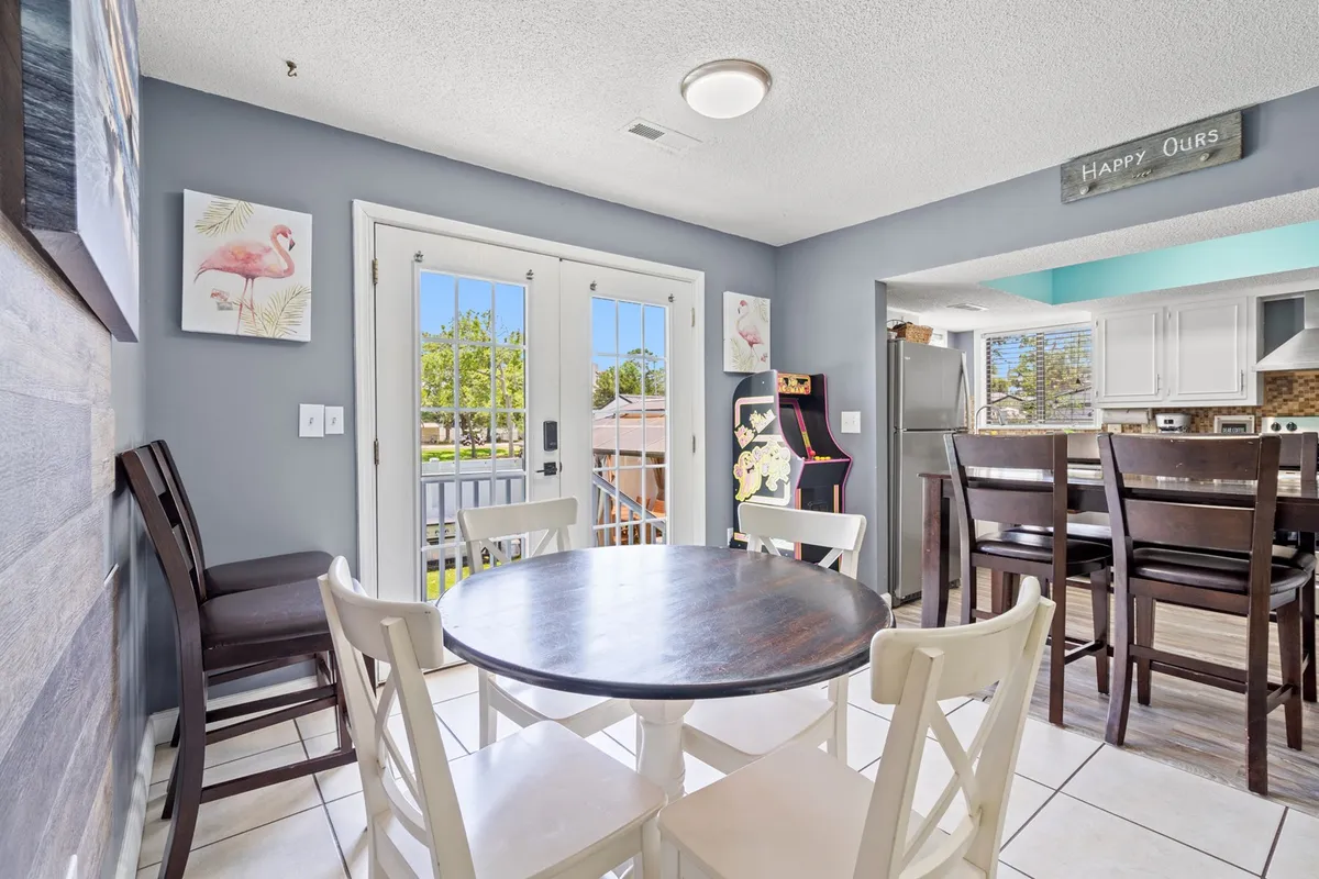 A bright and cheerful interior dining space featuring a round table, French doors that open to the outdoors, an arcade machine in the corner, and bar stools along the wall. The setting is designed for group meals, casual gatherings, and friendly games.