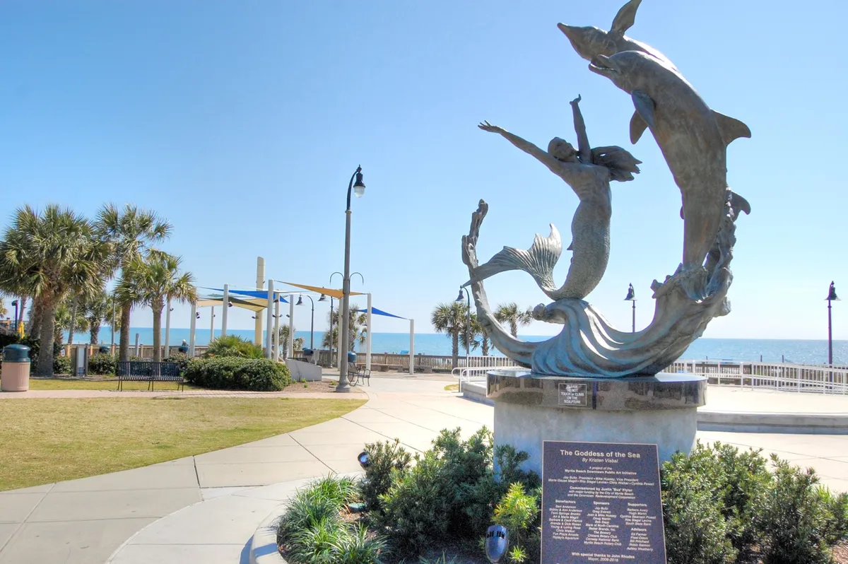 A joyful sculpture of a mermaid and dolphins sits along the oceanfront promenade, surrounded by swaying palm trees. The expansive blue ocean and sky stretch out behind, suggesting a vibrant mix of art, nature, and leisure activities available nearby.