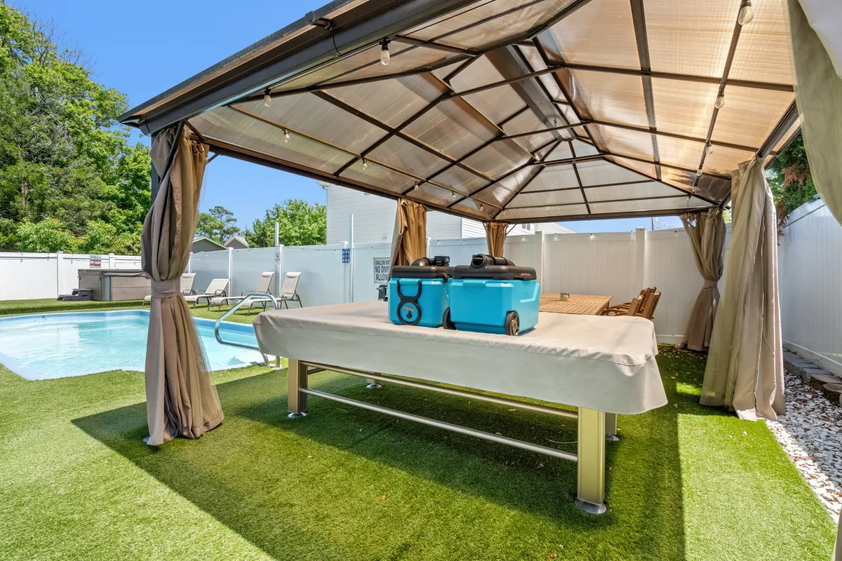 A sunlit backyard features a sparkling swimming pool surrounded by lounge chairs, a shaded gazebo for outdoor dining, and a beverage cooler nearby. The inviting space is set up for both relaxation and social gatherings, evoking a true resort atmosphere.