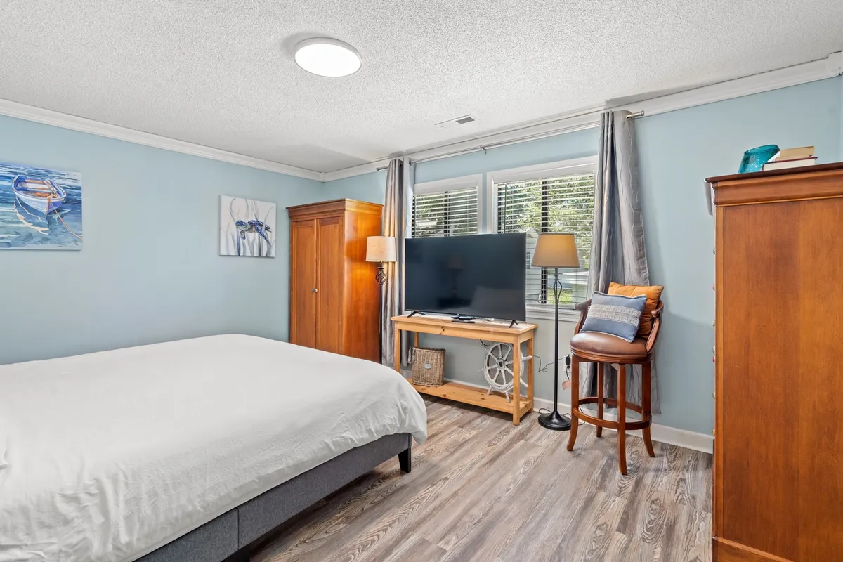 This bedroom features a bed with plush white bedding, decorative artwork above the headboard, wooden furniture, and a television unit with a lamp. Sunlight filters into the room, creating a calm and serene atmosphere ideal for unwinding after a busy day.