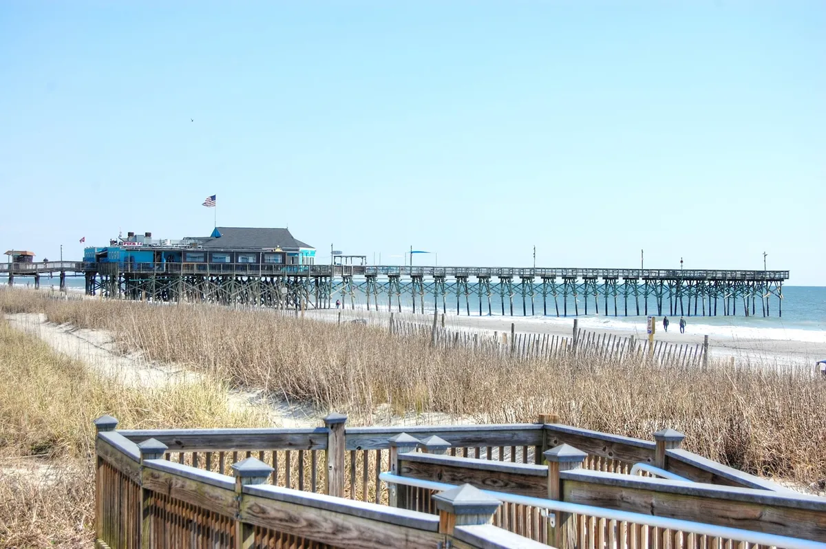 A tranquil view of a long pier stretching over the ocean, bordered by sand dunes and a boardwalk. The scene exudes a sense of adventure and relaxation, perfect for fishing, strolling, or simply enjoying scenic ocean views.