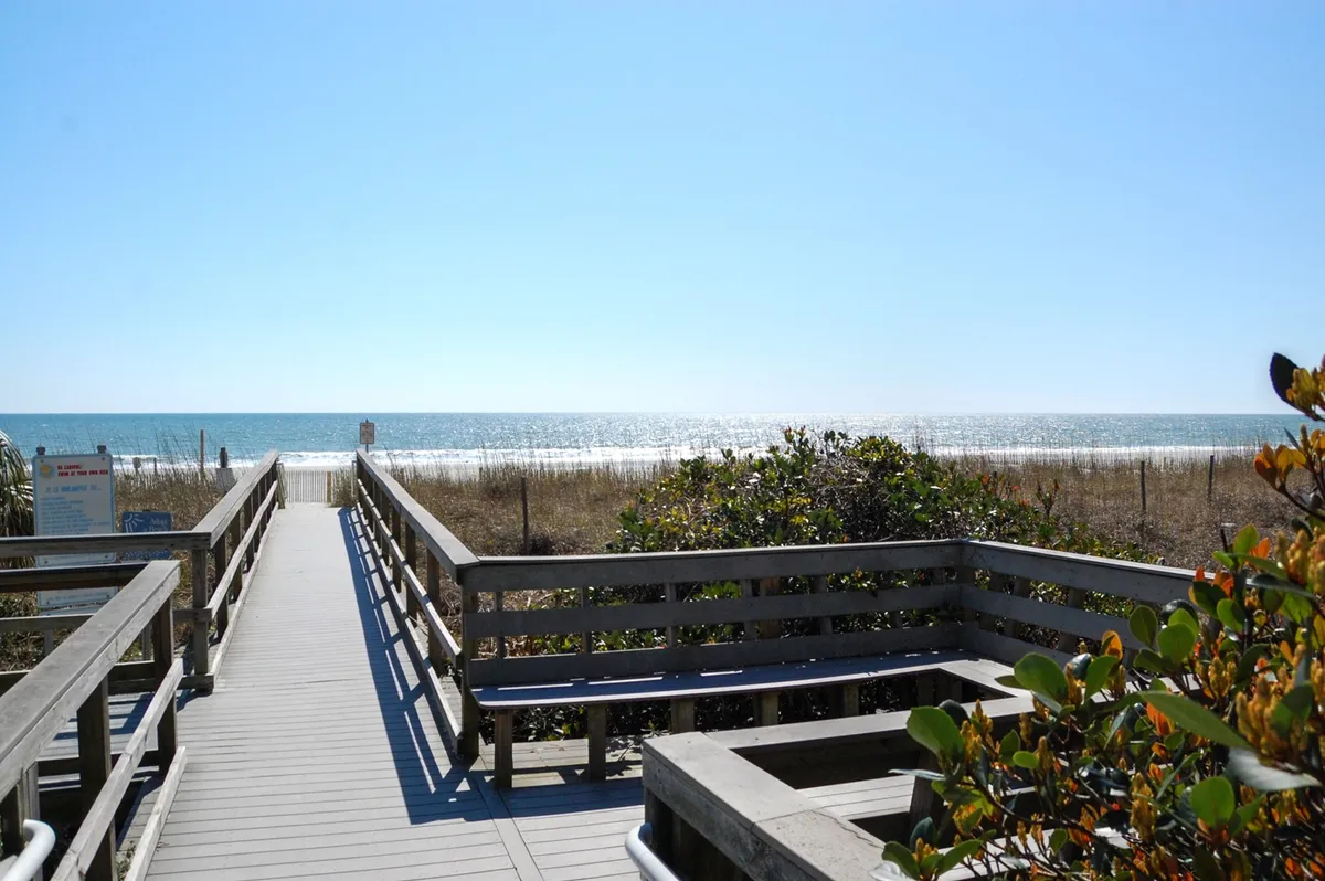 A sunlit wooden boardwalk leads across grassy dunes toward an expansive, sandy beach. The ocean glimmers in the background, with sunlight reflecting on the water and gentle waves rolling in. The scene conveys opportunity for morning walks, beachcombing, and the soothing ambiance of coastal living.