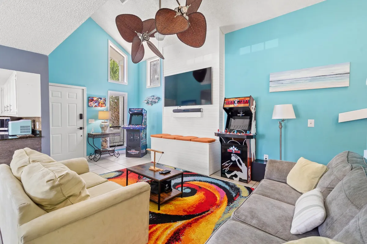 This vibrant interior scene showcases a spacious living area featuring a colorful rug, multiple stand-up arcade machines, and a large TV mounted on the wall. Comfortable seating and a ceiling fan contribute to a relaxed, welcoming environment, perfect for movie nights and game tournaments.