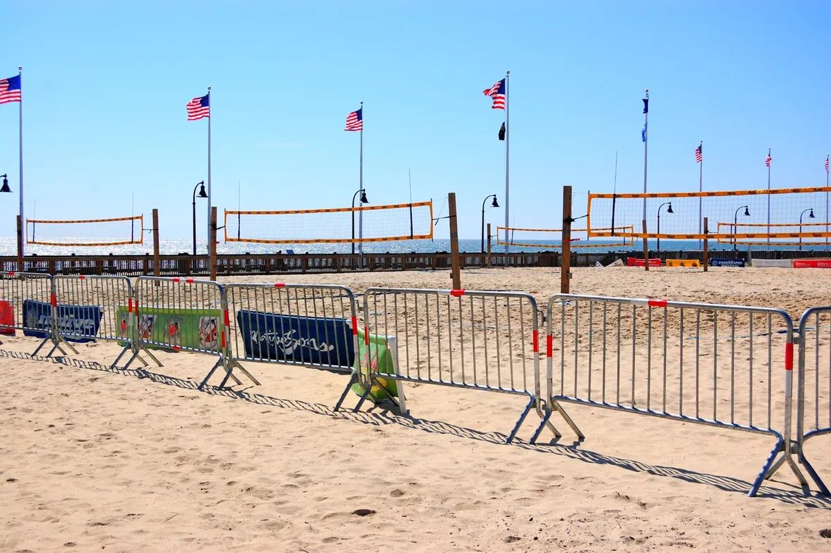 The scene shows lively beach volleyball courts with American flags in the background, bustling with energy and set against the backdrop of the open ocean. This lively, festive environment hints at social gatherings and active nightlife options along the beachfront.