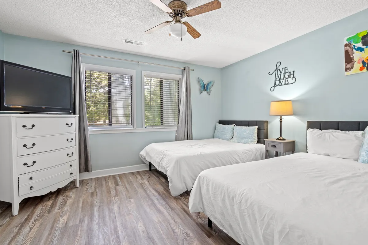 This image shows a bedroom with two beds dressed in crisp white bedding, a wall-mounted television, dressers with drawers, a decorative butterfly on the wall, and a lamp beside the bed. The room exudes comfort and is designed for both relaxation and shared moments, making it perfect for groups or families.