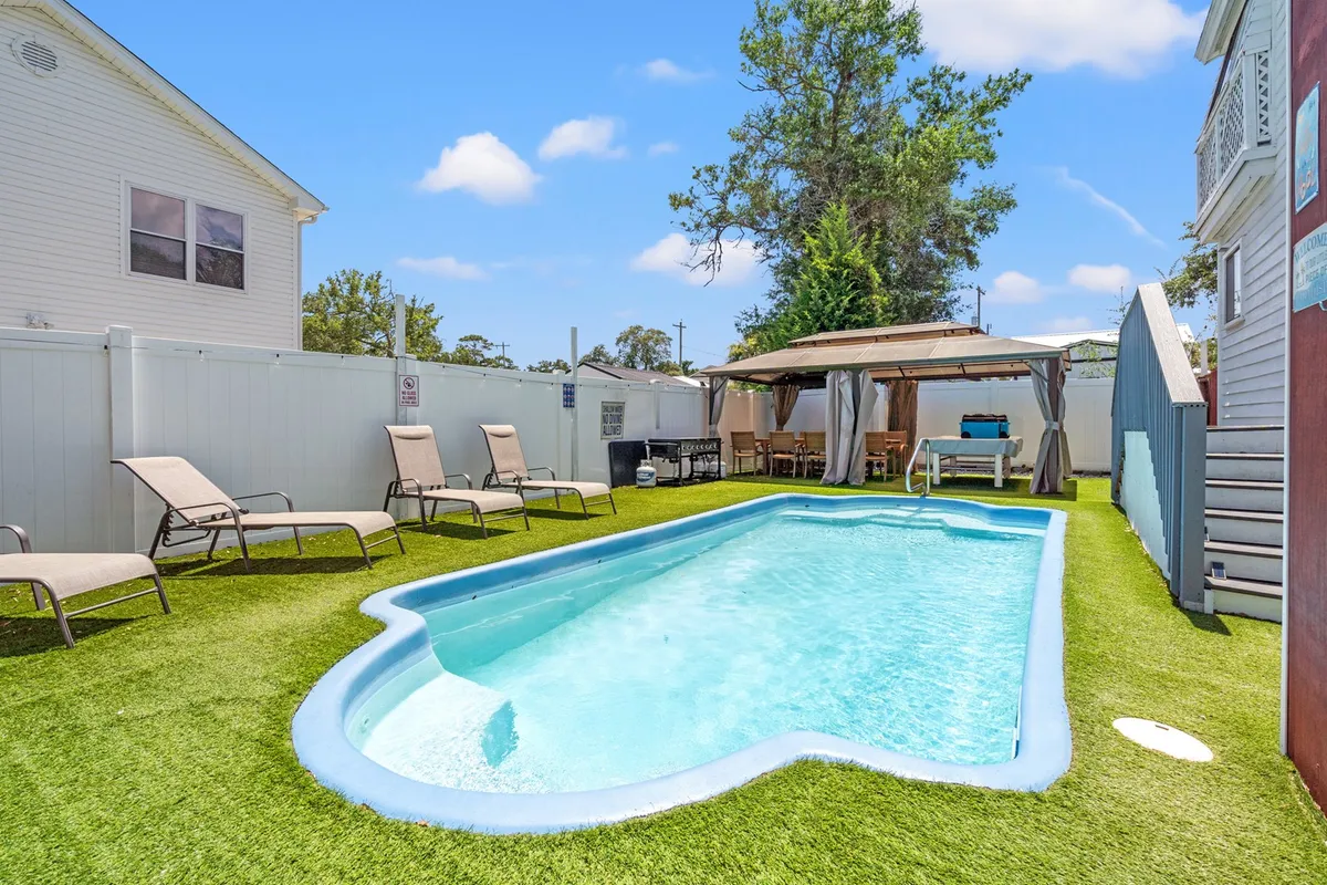 The image showcases the sun-kissed backyard of a spacious single-family home, with a sparkling swimming pool as the centerpiece. Surrounding the pool are inviting lounge chairs and a stylish cabana, all set amid lush green grass—perfect for relaxing, outdoor dining, or soaking up the cheerful North Myrtle Beach vibes.