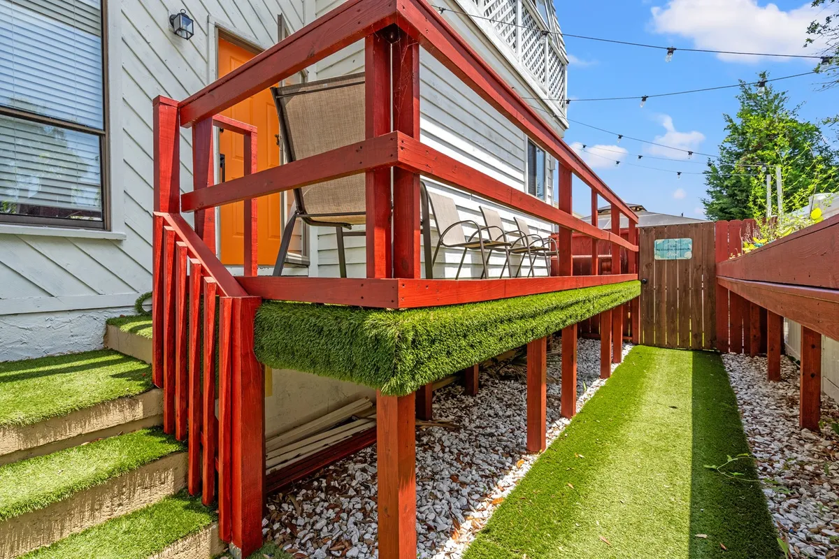 A cheerful backyard setting features a bright orange door, a modern red railing, and a plush grass-like seating area perfect for outdoor dining or casual gatherings. The space is designed for comfort and entertaining.