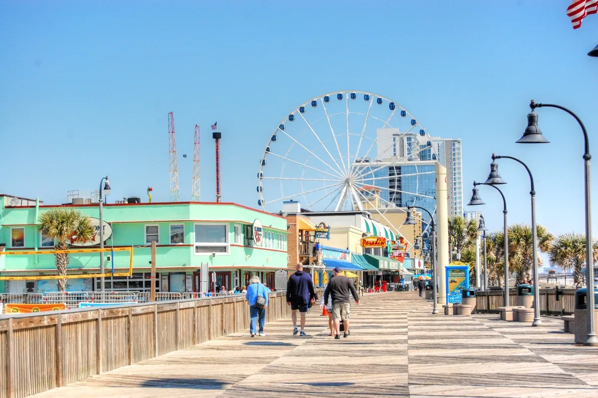 A vibrant scene showcasing a ferris wheel along a lively boardwalk, with colorful buildings, people enjoying the area, and palm trees setting a coastal vibe. The energy of the scene captures the excitement and variety of entertainment options available for families near the beach.