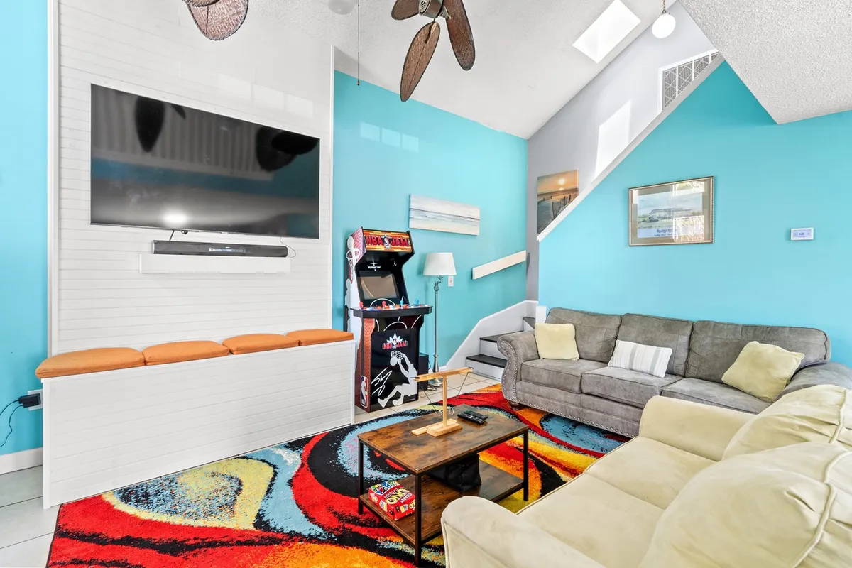 The image highlights a large flat-screen TV, a vibrant area rug, a classic arcade game machine, and a comfortable seating arrangement. The setup is clearly intended for socializing, group activities, and family gatherings, with a casual and fun atmosphere.