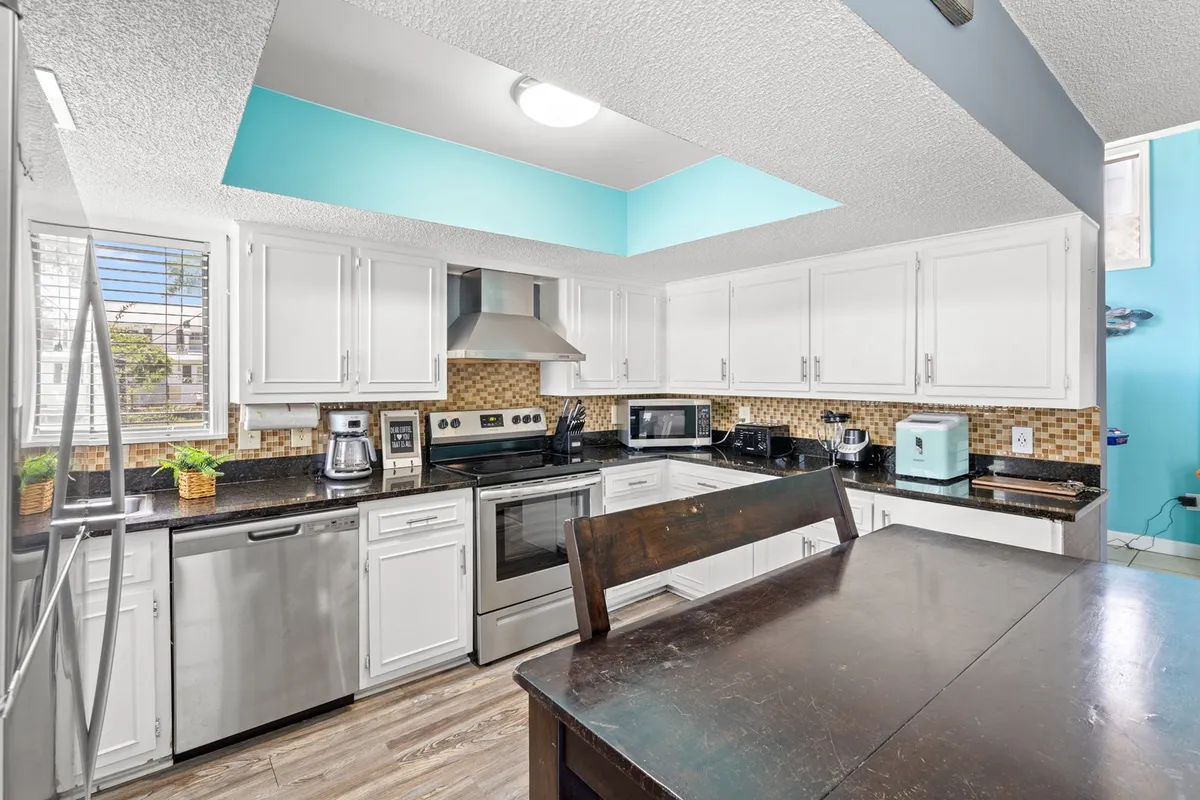 A contemporary kitchen scene centered on stainless steel appliances and a granite countertop. The adjoining wood dining table seats multiple people, reflecting the promise of group meals, brunches, and casual get-togethers. Subtle details like the light blue ceiling add a fresh, inviting touch.