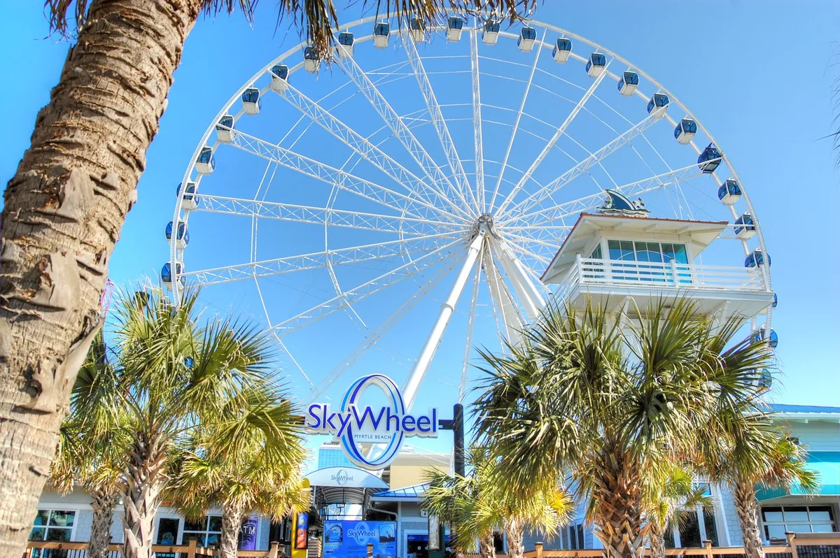 A bright exterior shot focused on the towering SkyWheel, framed by palm trees and an inviting entrance. Families and visitors gather at the base, hinting at the excitement of soaring above the coast and taking in panoramic views.