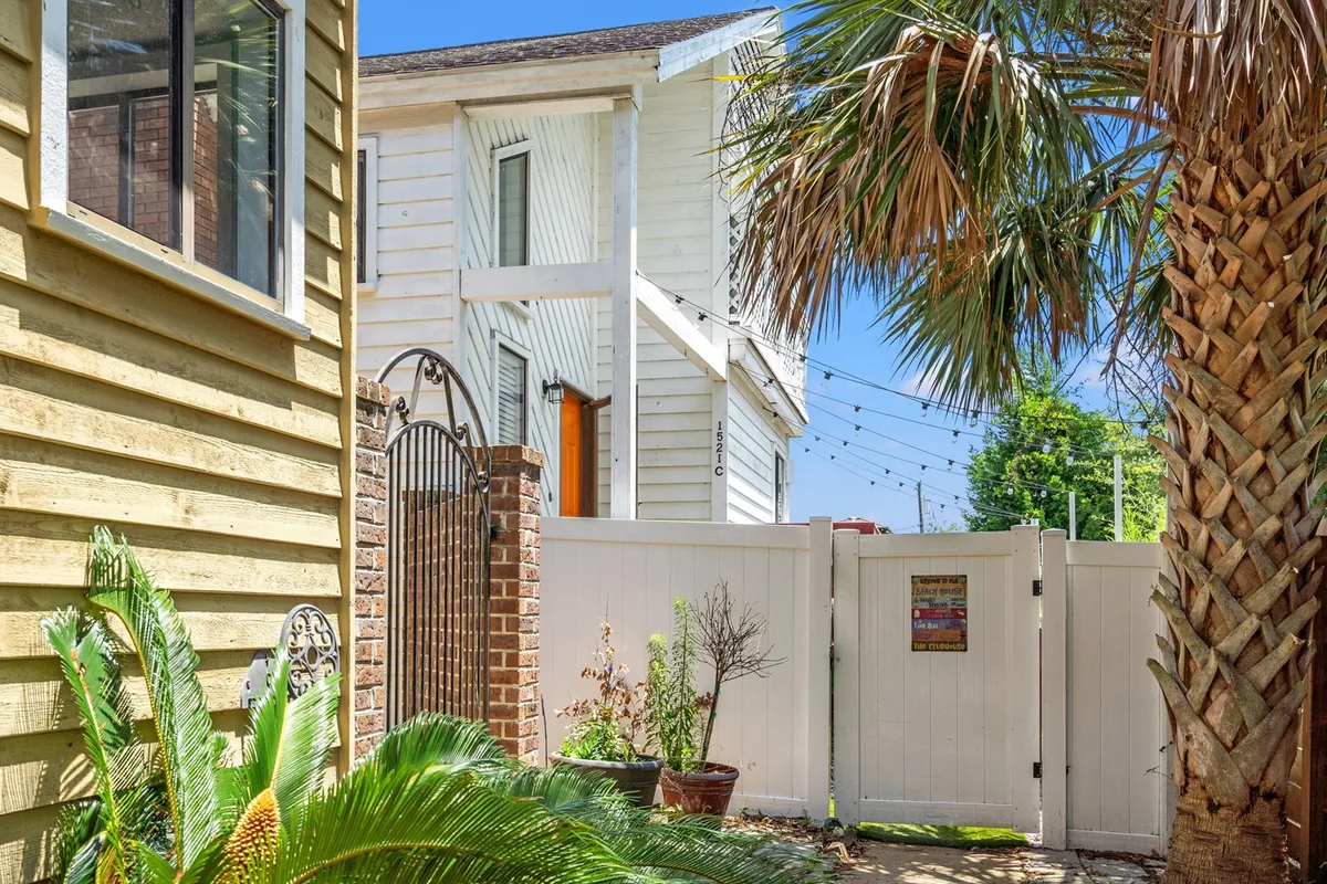 A welcoming view of a white wooden house framed by a brick gate, with lush green plants and tall palm trees surrounding the entrance. The yard is spacious and inviting, ideal for outdoor gatherings or group celebrations.